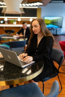 Sarışın kadın laptopta çalışıyor. Ofis çalışma alanında masada oturuyor. Erkek iş arkadaşı yakınlarda iş planlayarak zaman geçirir.