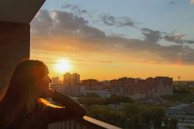 Uzun saçlı kadın balkondan akşam şehrini geziyor. Bayan gün batımında aydınlanan yüksek binalara bakmayı ve onları keşfetmeyi seviyor.