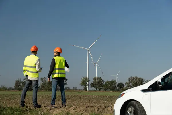 风车在农村为电动汽车发电 戴橙色头盔的风力涡轮机技师站在强大的风车旁边 免版税图库照片