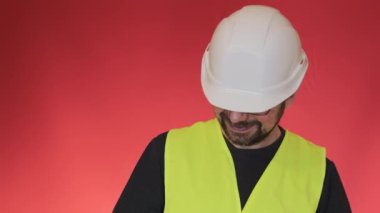 Gözlüklü olumlu müteahhit başını kaldırıp kameraya gülümsüyor. Beyaz kasklı ve yelekli sakallı adam kırmızı arka planda duruyor.