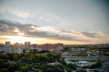 Gün batımı beyaz bulutların arasından doğar ve işlek şehri aydınlatır. Yol üzerinde çok katlı binalar yükseliyor ve etrafı karanlık ağaçlarla çevrili.