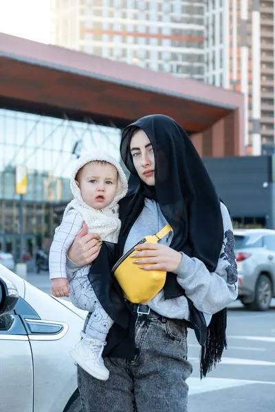 英国Kyiv 2021年9月27日 身穿黑色头巾的妇女抱着孩子 靠着繁忙的停车场 享受照看幼儿的乐趣 可爱的儿子高兴地环顾四周 免版税图库图片