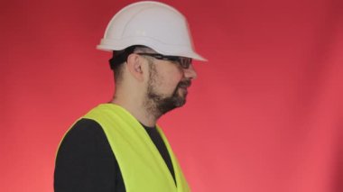Beyaz kasklı profesyonel mühendis ekipmanları gösteriyor. Üniformalı sakallı adam stüdyoda kırmızı arka planda duruyor.