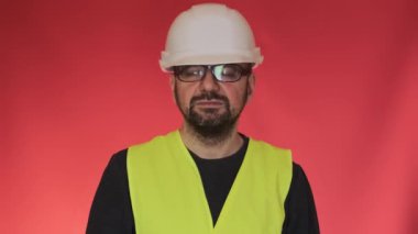 Koruyucu başlık takan ciddi mühendis kameraya dönerek aşağı bakıyor. Kırmızı arka planda üniformalı sakallı işçinin portresi.