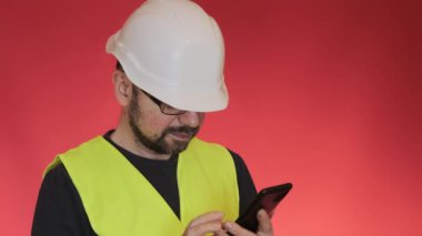 Beyaz kasklı sakallı mühendis akıllı telefondan proje planına bakıyor. Üniformalı işçiler kırmızı arkaplanda duran bir cihaz kullanıyorlar.