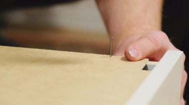 Erkek eli çivi kafasına çekiçle vurur. Prefabrik mobilyaların ahşap parçalarını tutturur. Tamirci, beyaz kontrplaktan mobilyaları dikkatlice monte eder.