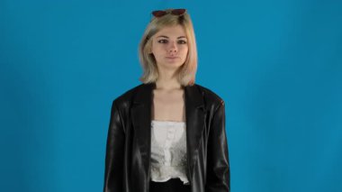 Sarışın kadın, mavi arka planda el hareketleriyle takip etmek için davet ediyor. Deri ceketli ve güneş gözlüklü genç bir kadın şakacı bir şekilde flört ediyor.