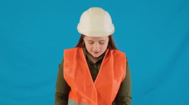 Sorumlu bayan müteahhit mavi arka planda koruyucu eldivenleri ayarlar. Üniformalı genç kadın güvenlik standartlarını korumak için çalışmaya hazırlanıyor.
