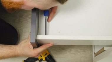 Elinde tornavida olan bir zanaatkar çekmeceyi bitirir. Sakallı uzman, düz zeminde oturan küçük elementleri vidalıyor ve saat mekanizması çalışıyor.