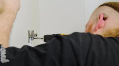 İşitme cihazı olan tamirci kapı menteşesini yüzeye çıkarır. Sakallı zanaatkar elinde tornavida tutuyor ve iyi aydınlatılmış odada mobilya montajında çalışan nesneleri tamir ediyor.