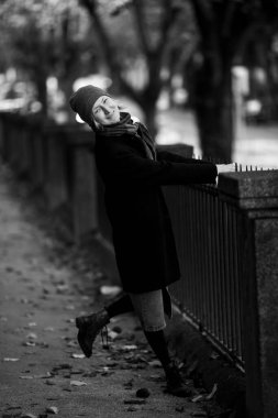 Park çitlerinin yanında poz veren sonbahar montlu bir kadın. Siyah beyaz fotoğraf.