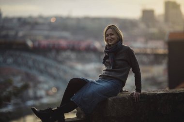 Porto, Portekiz 'de, alacakaranlıkta antik bir taş duvarın yanında oturan bir kadın. Arka planda, köprünün bulanıklığında Dom Luis.