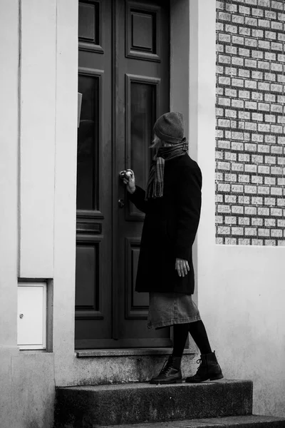 A woman on the porch of a house knocks on the door. Black and white photo.