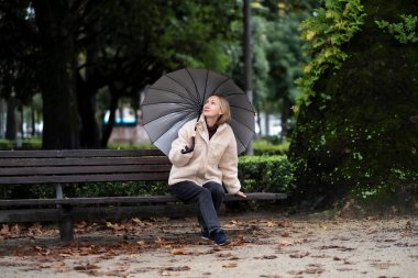 A woman with an open umbrella sits on a bench in the Park.