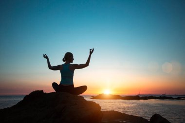 A woman does yoga, meditating by the ocean during sunset.   