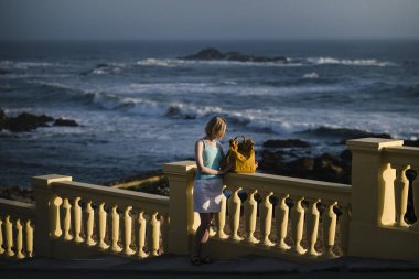 A woman with a yellow backpack stands on the Atlantic seafront, Porto, Portugal. 