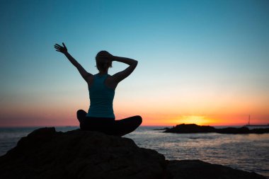 Silhouette of a woman in lotus pose doing yoga on the oceanfront during a sunset.   