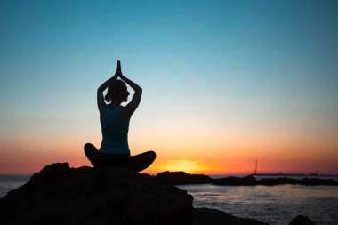 A silhouette of a middle-aged woman doing yoga on the ocean, seeing off the sun.   
