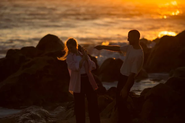 驚くべき日没の間に岩の多い海洋前線をうろうろする若いカップル — ストック写真