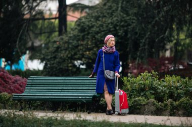 Elinde bavul olan bir kadın bankın yanında bekliyor..