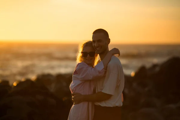 黄金の夕日の間に岩の多い海の海岸に抱擁する若いカップル — ストック写真