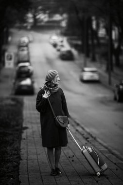 Elinde bavul olan bir kadın yolun kenarında duruyor. Siyah beyaz fotoğraf.
