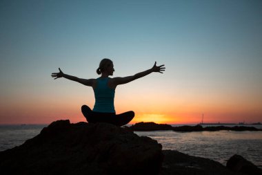 Gün batımında deniz kenarında oturan yoga yapan bir kadının silueti..