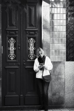 Eski bir Portekiz şehrinin caddesinde akıllı telefonuyla yazı yazan bir kadın. Siyah beyaz fotoğraf.