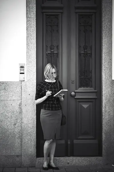 A woman with a notebook and pen stands outside a house. Black and white photo.