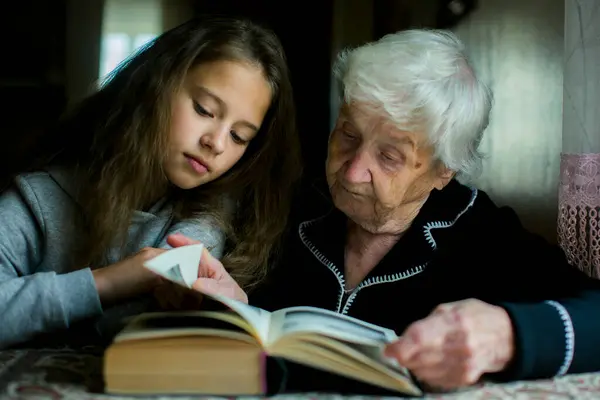 An old woman is reading a book with her granddaughter.