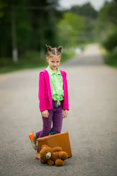 道端では 明るい服装で飾られた少女は 発見の興奮を象徴する彼女のスーツケースと最愛のプラッシュおもちゃを運びます ロイヤリティフリーのストック写真