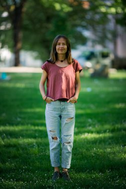 Tişörtlü ve yırtık kotlu genç bir kadın parkta dikiliyor, uzaktan derin derin bakıyor..