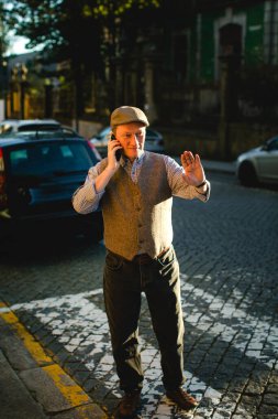 Sokakta dikilen, cep telefonuyla konuşan, kep, kot pantolon ve yelek giyen bir adam..