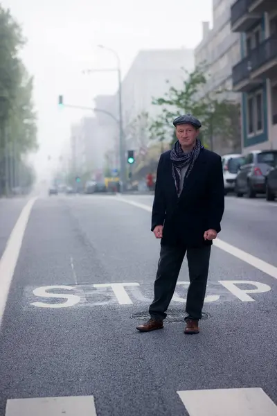 A man wearing a jacket, cap, and scarf is standing on the urban street on a cool morning.