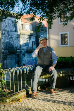 Orta yaşlı bir adam parkta otururken telefonla konuşuyormuş gibi yapar. Huzurlu ortama biraz neşe katar..