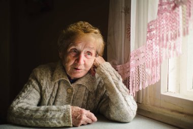 Süveter giymiş yaşlı bir kadın pencerenin kenarında oturur, sakin bir ifadeyle dışarı bakar, huzurlu bir yansıma anı yakalar..