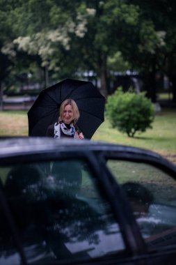 Bir araba yaklaşıyor ve şemsiyeli bir kadının yanında duruyor..