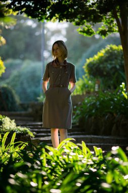 Bir kadın, eski bir parktaki yaprakların arasından süzülen noktalı ışıkta duruyor..