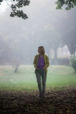 Sis ve yumuşak ışıkla çevrili sisli bir şehir parkında kot pantolon ve leylak renkli bir kazak giyen bir kadın, sakin ve huzurlu bir atmosfer yaratıyor..