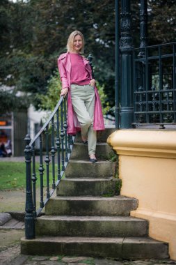 Pembe bir palto giymiş bir kadın, serin bir sonbahar havası ve sakin bir sonbahar havasıyla çevrili bir parkın taş basamaklarında duruyor..