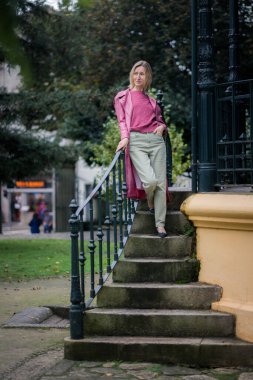 Pembe ceketli bir kadın parktaki taş basamaklarda hareketsiz durur, sonbahar soğuğu dingin ve yansıtıcı atmosfere katar..