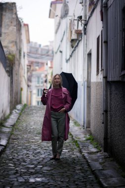 Pembe paltolu bir kadın dar bir sokağın kaldırım taşı sokağında duruyor, elinde şemsiyeyle bulutlu gökyüzü yağmuru ima ediyor, kasvetli ve sessiz bir sahne yaratıyor..