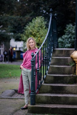 Sessiz bir parkta, taşın yanında duran pembe ceketli bir kadın serin bir sonbahar gününün sükunetini kucaklıyor..