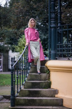 Pembe paltolu bir kadın sonbaharda parktaki taş basamaklarda duruyor, bulutlu hava sakin ve yansıtıcı bir atmosfer ekliyor..