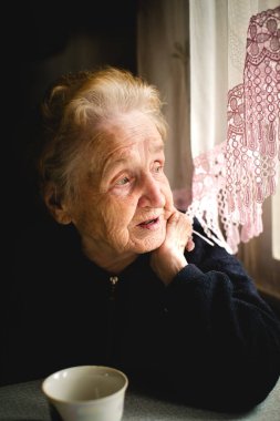 Yaşlı bir kadın düşünceli bir şekilde pencereden dışarı bakar, kendi yansımasında kaybolur..