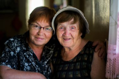 Yaşlı bir kadın, yetişkin, orta yaşlı kızıyla fotoğraf çektirmek için poz veriyor. İkisi de sıcak bir şekilde gülümsüyor ve güzel bir aile bağı kuruyor..