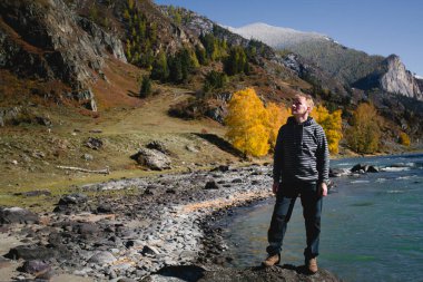 Bir erkek gezgin, Altai Cumhuriyeti 'ndeki bir dağ nehrinin kıyısında duruyor. Hızla akan suya ve engebeli manzaraya bakıyor..