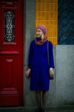 Orta yaşlı bir kadın, leylak paltolu, bereli ve boyun atkısı Portekiz 'deki bir evin uzun kapısının yanında duruyor, şık görünüyor..