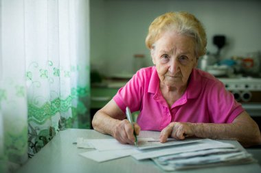 An elderly woman sits in her kitchen, filling out a stack of utility bills, her expression focused as she carefully manages her household expenses. clipart