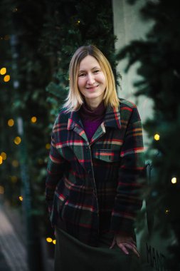 Kareli ceketli gülümseyen bir kadının portresi, köknar dalları ve arka planda yanıp sönen ışıklarla, sıcak bir tatil atmosferi yaratıyor..